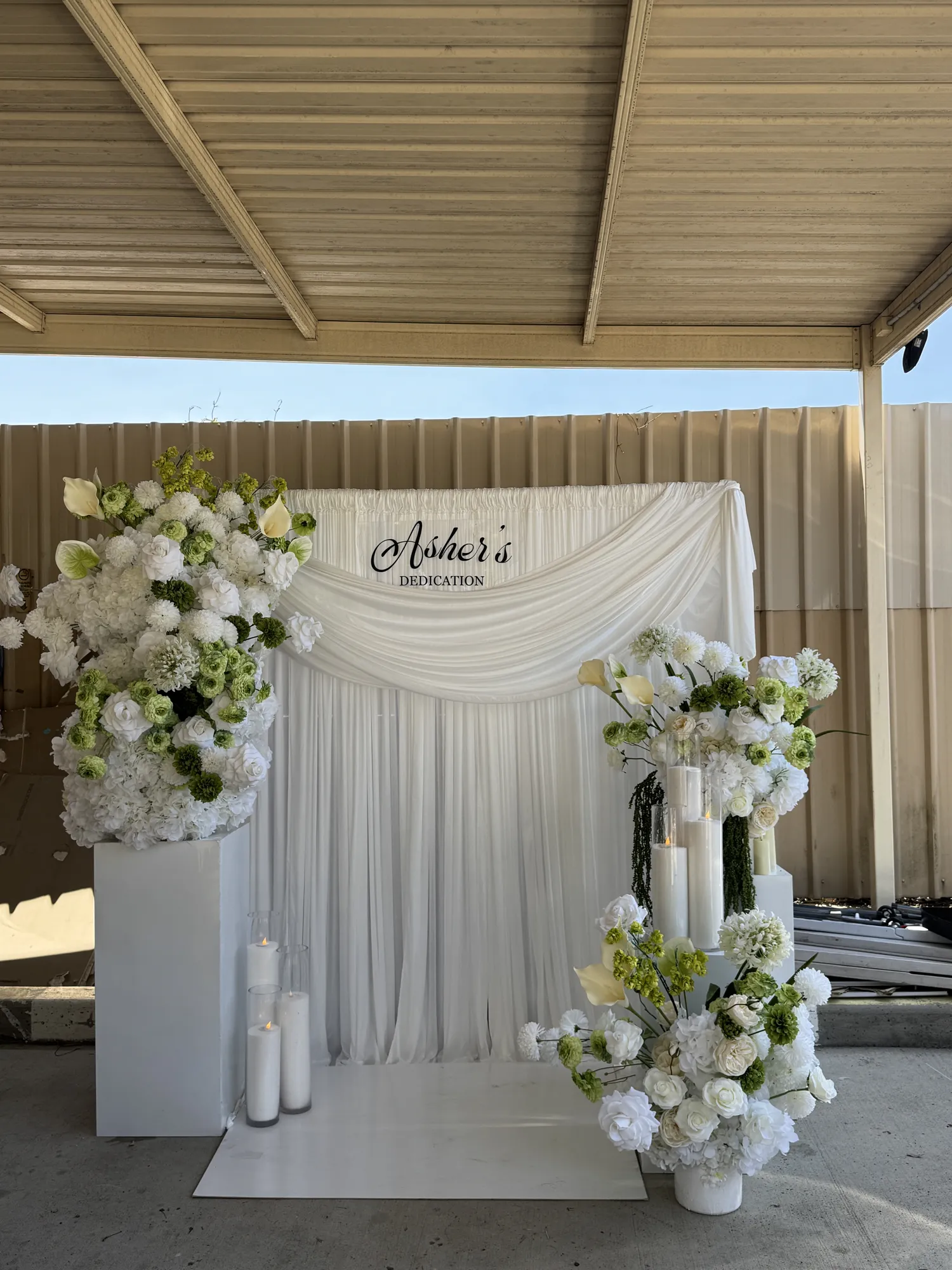A white and green floral backdrop with a dedication sign, seen from the right, with white flowers and lit candles.