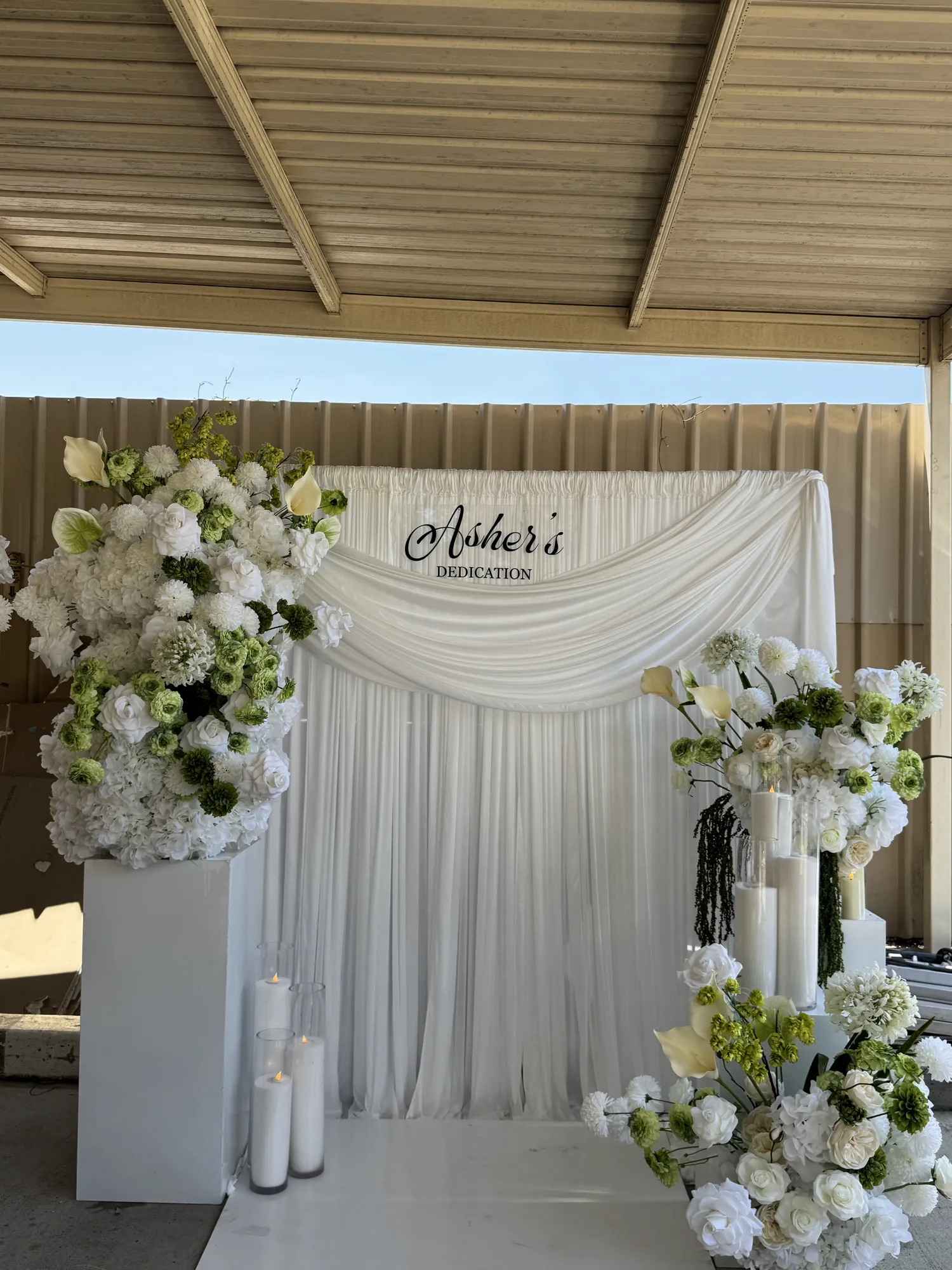 A white and green floral backdrop with a dedication sign, seen from farther right, with white flowers and lit candles.