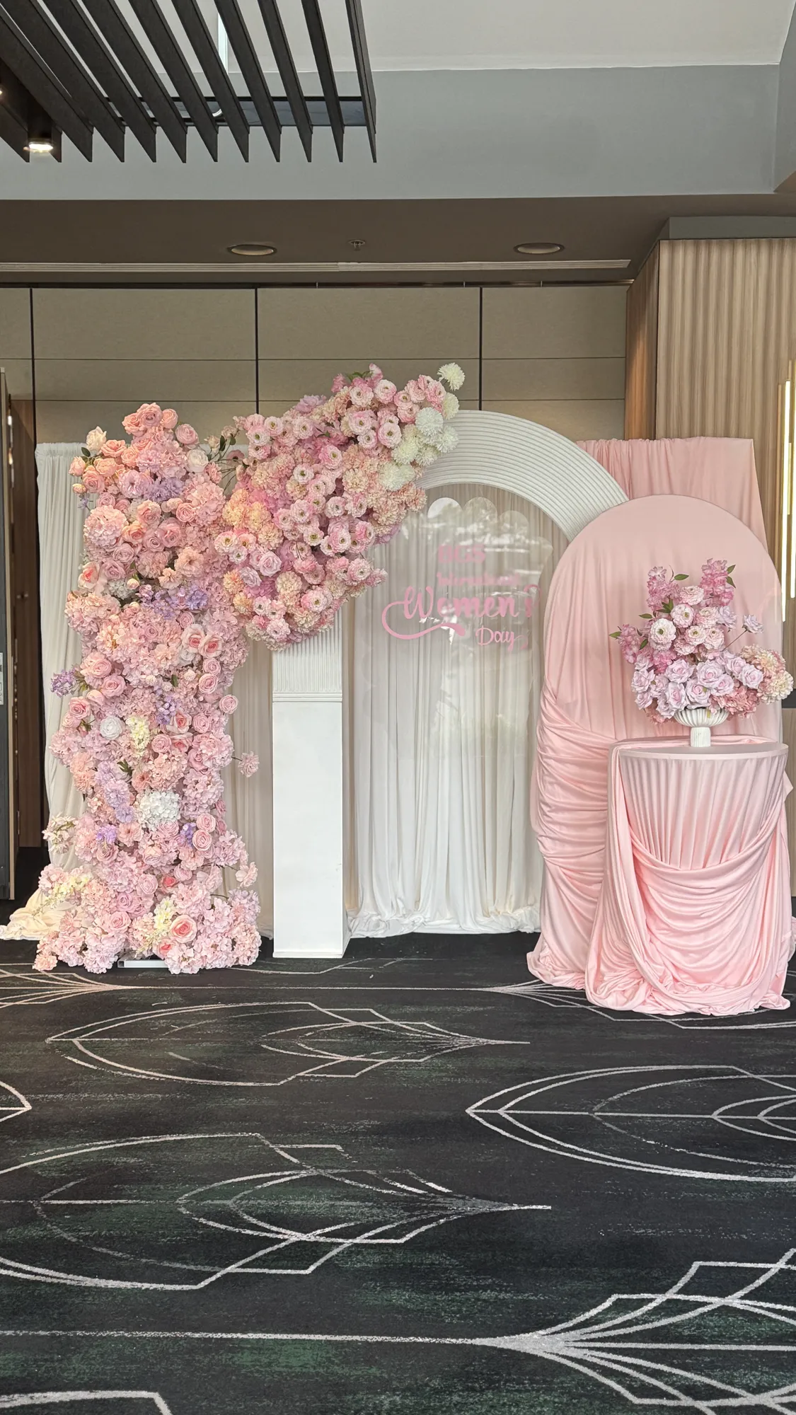 A white pedestal with a glass vase of pink and white flowers, a candle, and a small pink neon sign.