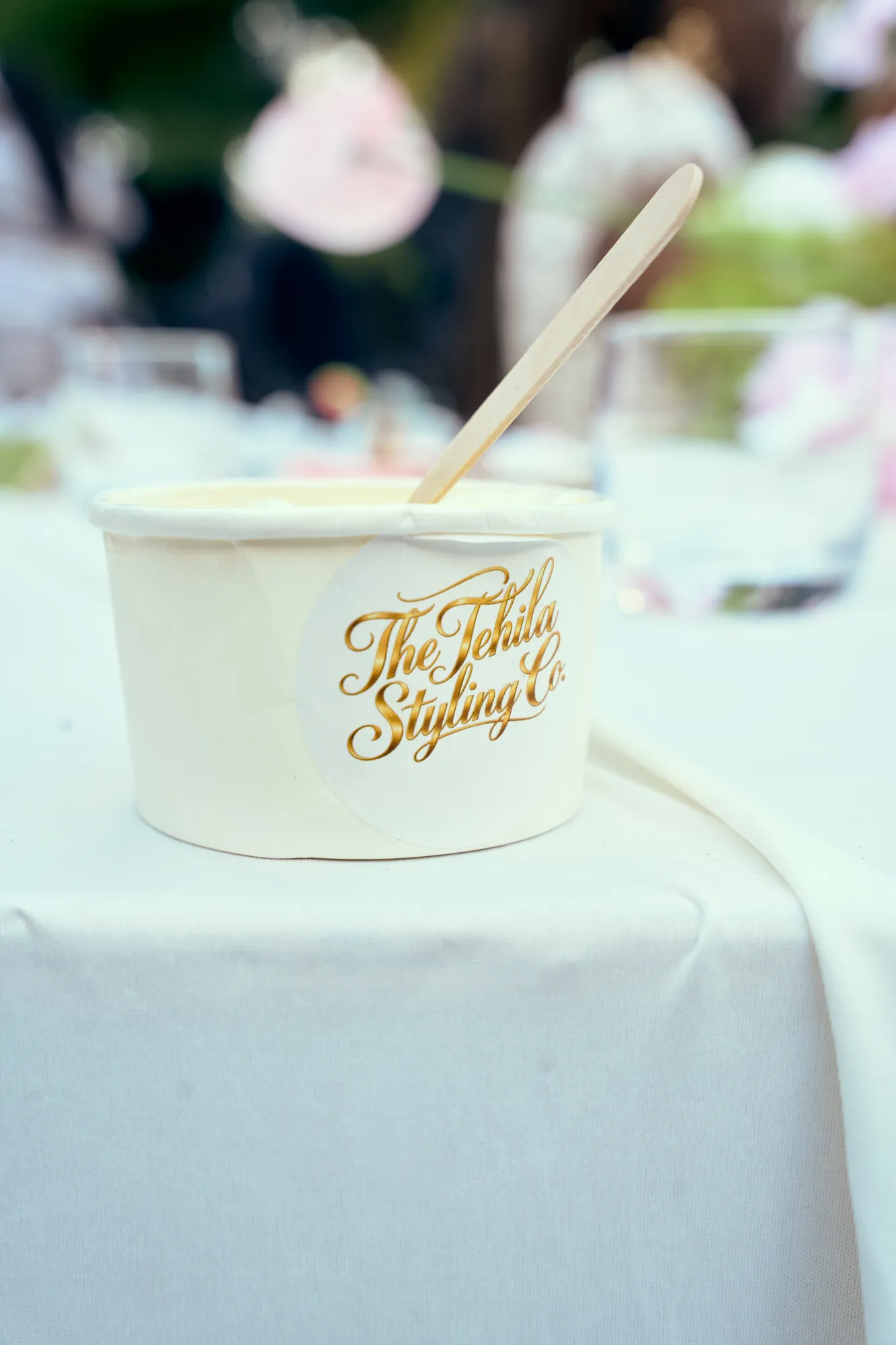 A hand holding a cup of chocolate gelato with a Tehila Styling Co sticker, beside pink and brown macarons.