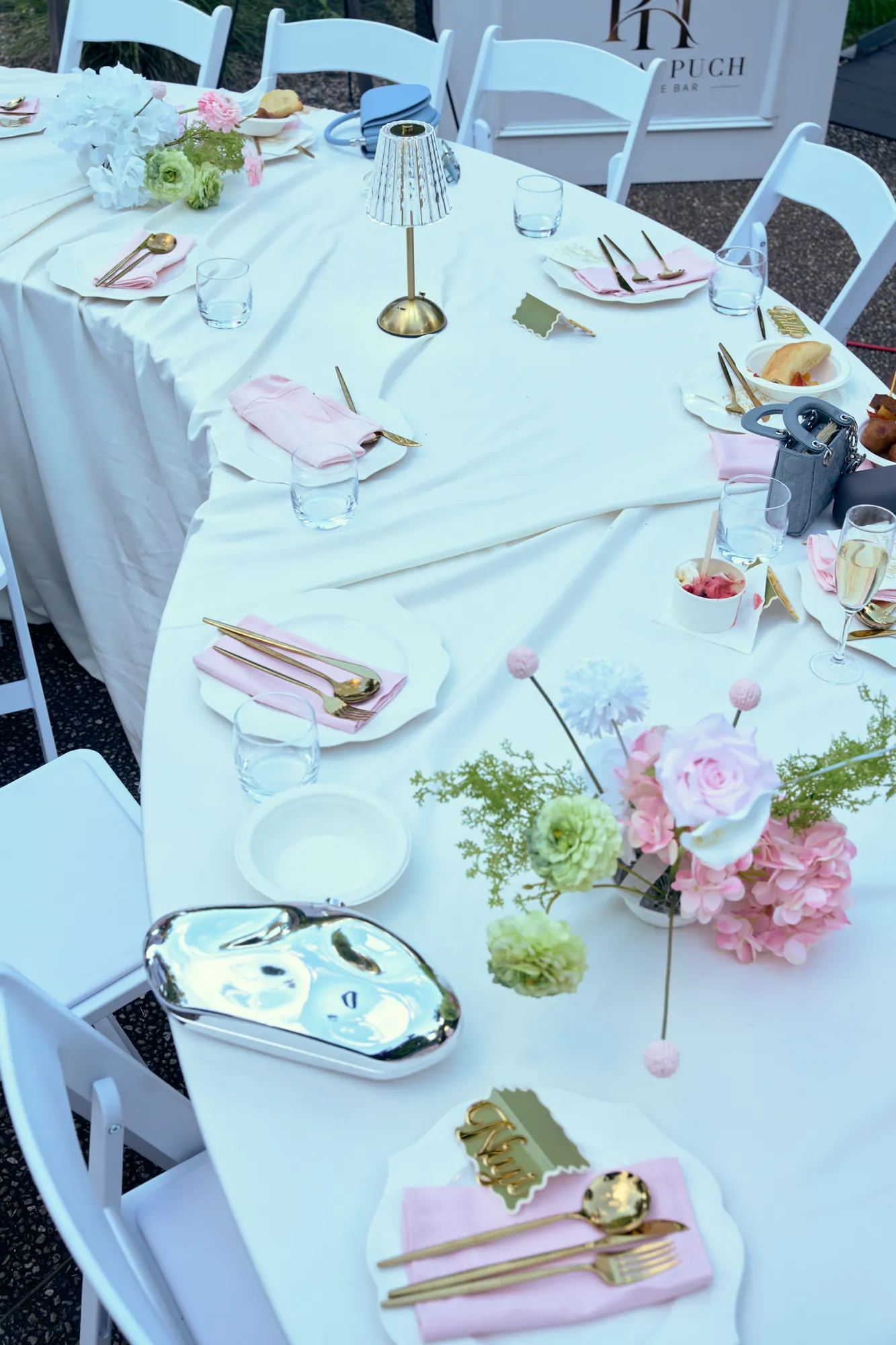 A long table in white linen set with pink napkins, gold cutlery, and low pink and white flowers.