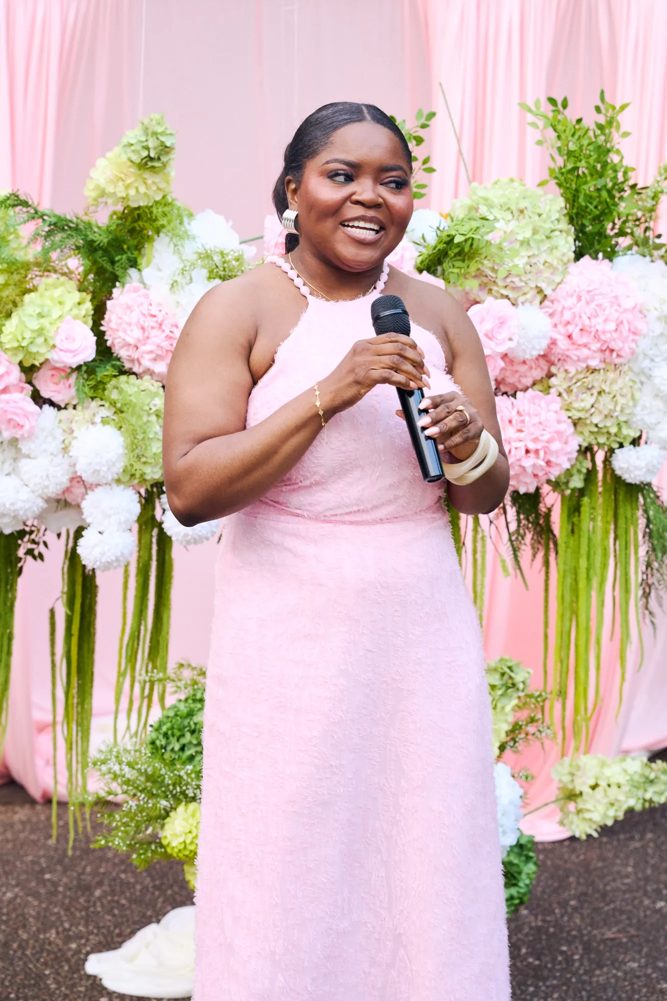 A host speaking into a microphone in front of a white floral backdrop and a pink neon sign.