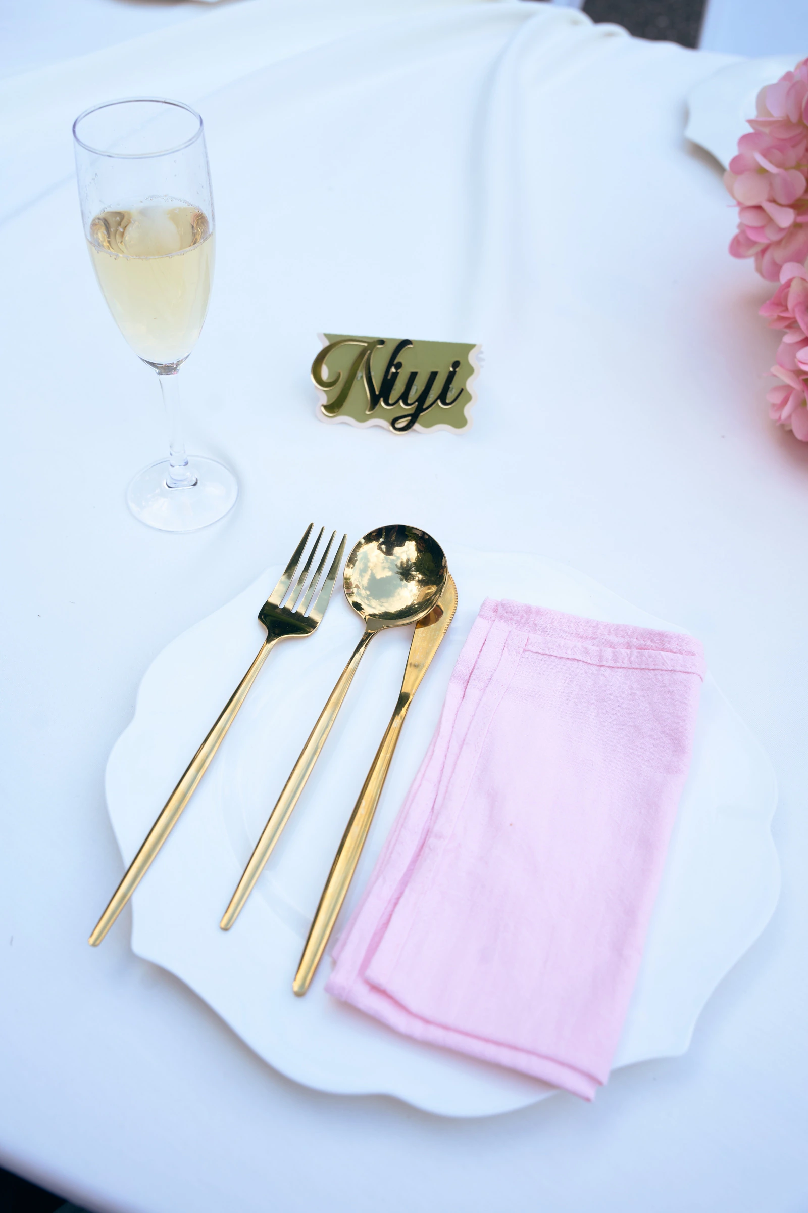 A place setting with a pink napkin, gold cutlery, a champagne flute, and a gold name card reading Niyi.