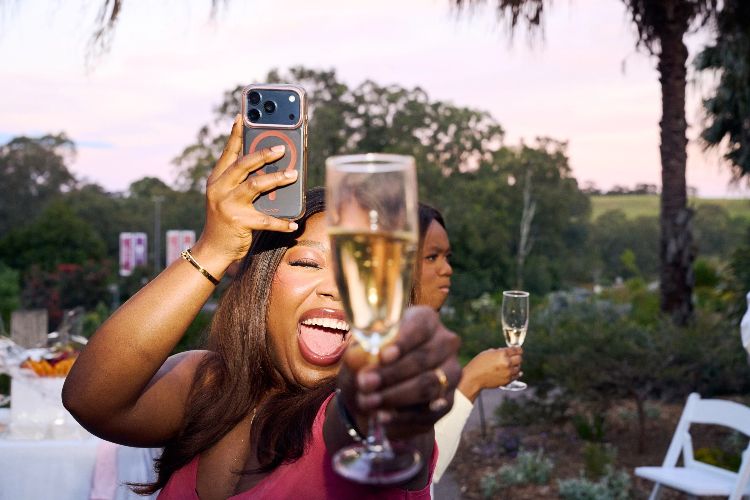A woman in a pink dress laughing at sunset, holding a phone and a glass of champagne toward the camera, with another guest holding a second glass behind her.