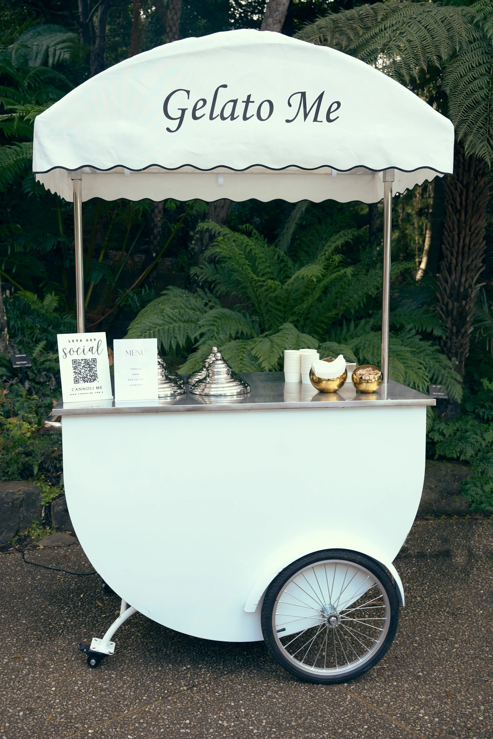 A white Gelato Me cart with a scalloped awning, set among tree ferns, with silver covers and gold cups on the counter.