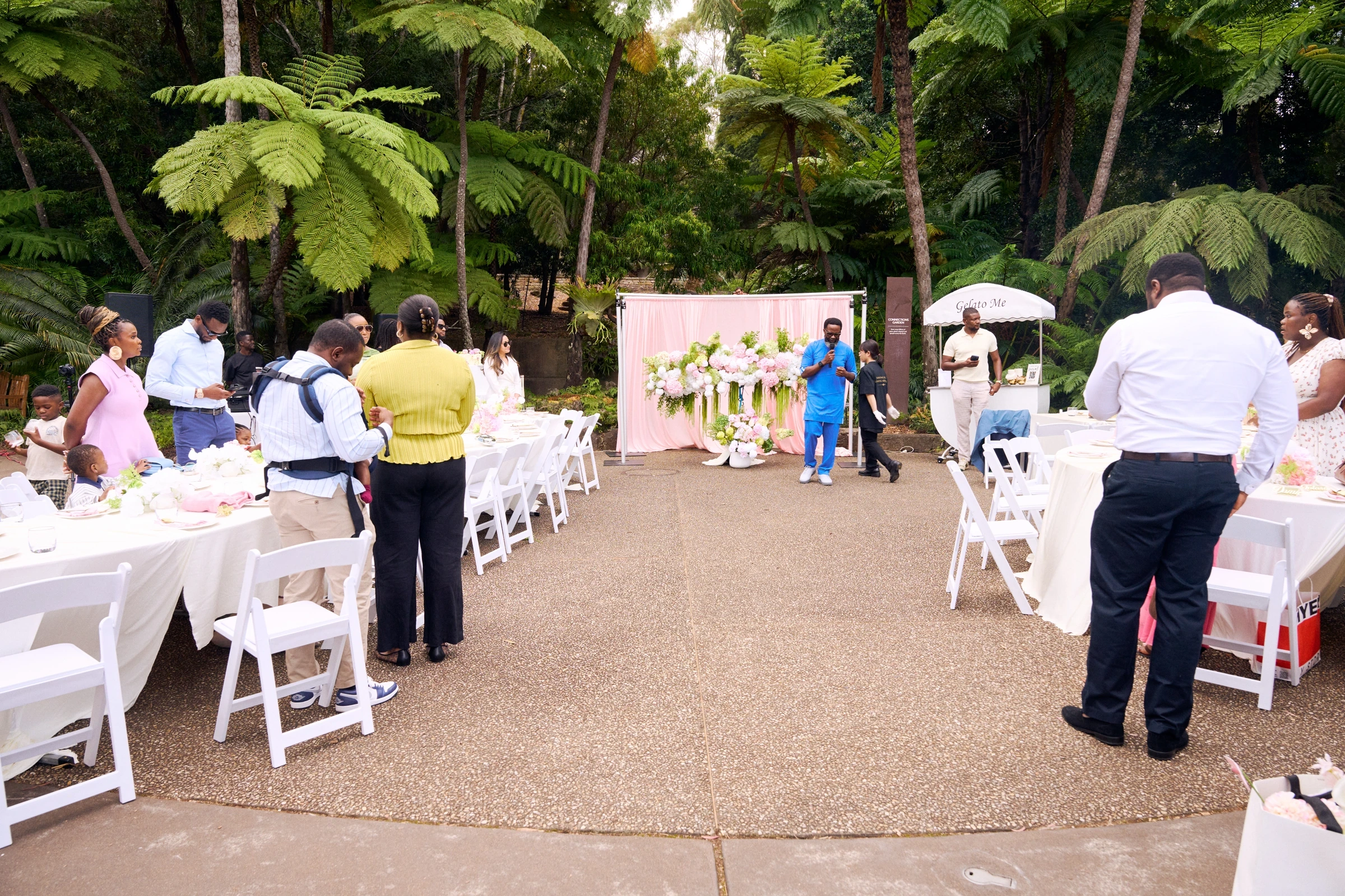 A garden venue under tree ferns, with long tables and white chairs, and a pink draped backdrop behind a pink and white floral installation.