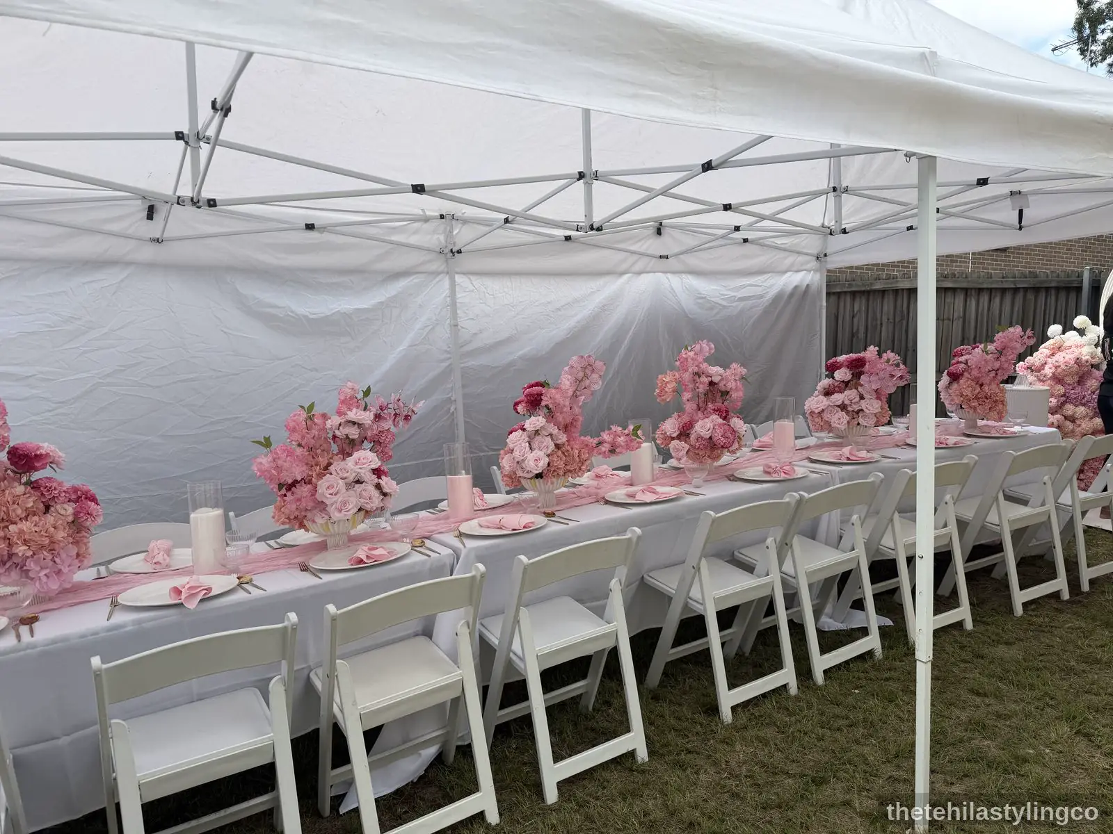 Curved outdoor banquet table in white linen, set with pink napkins, gold cutlery, and a pink and white floral centrepiece.