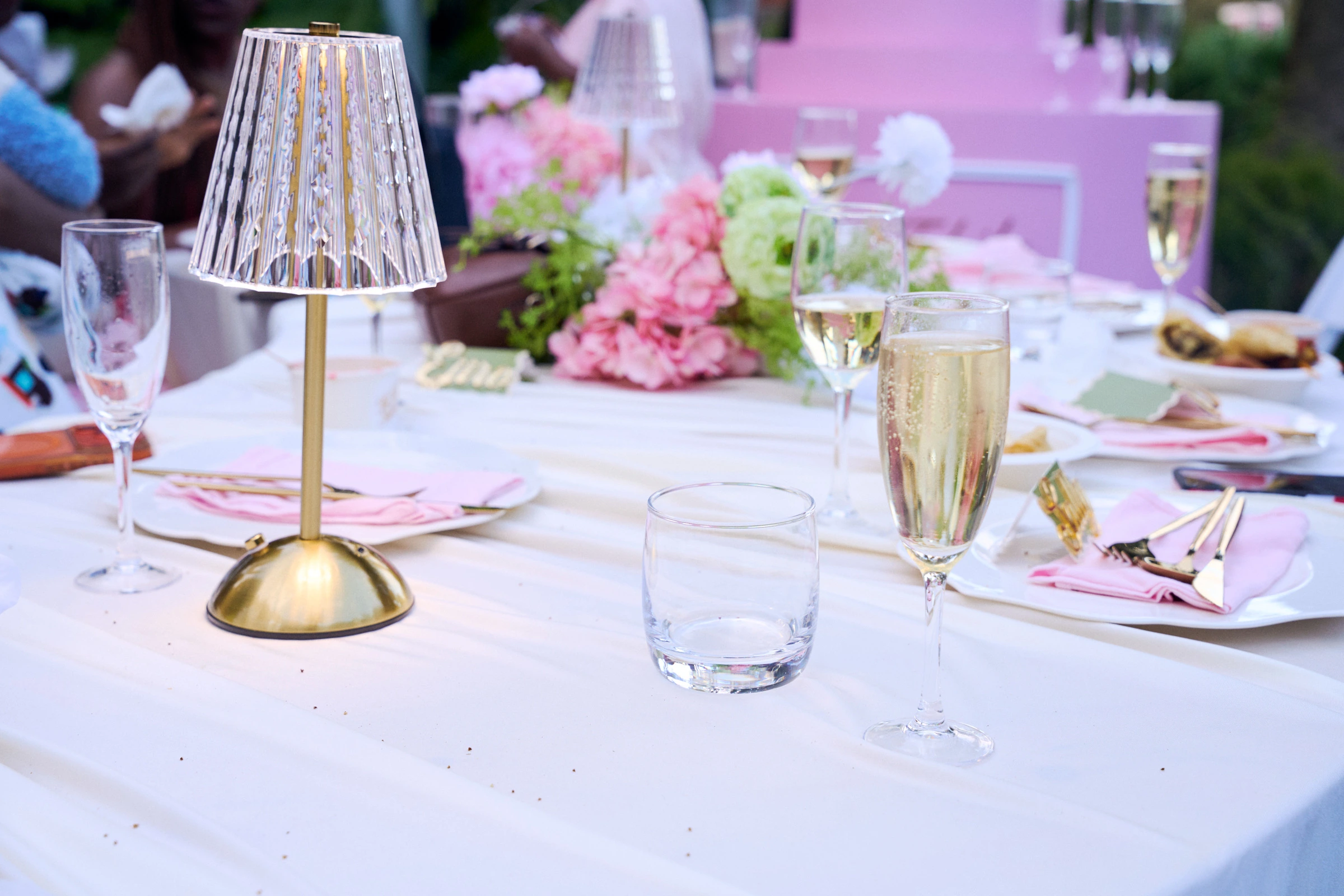A gold crystal lamp on white linen, with champagne flutes, a water glass, pink napkins, and gold cutlery, and pink and green flowers behind.
