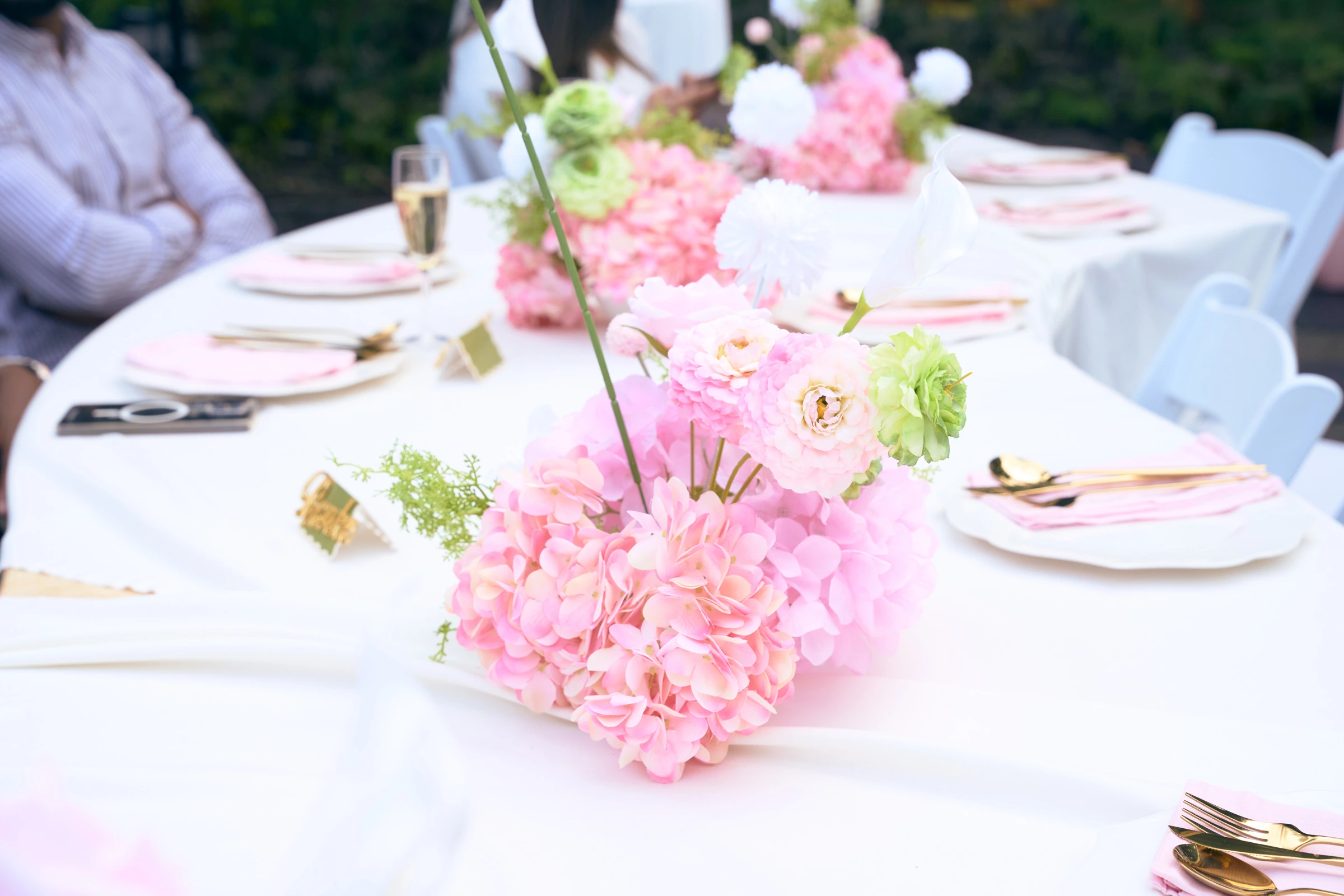 A low centrepiece of pink and white hydrangea, roses, and a calla lily on a round table in white linen, with gold cutlery, pink napkins, and gold place cards.