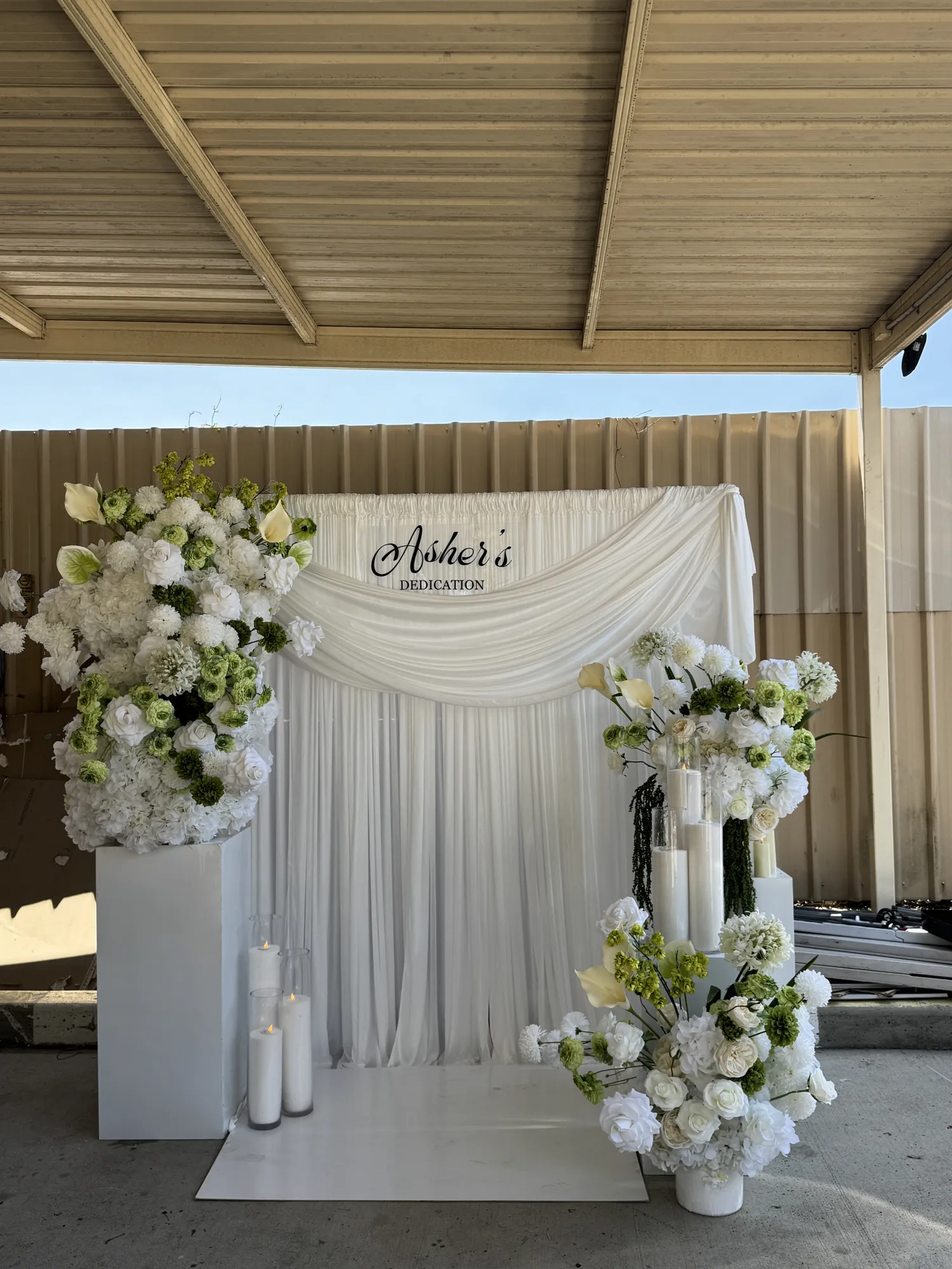 A white and green floral backdrop with a dedication sign, white flowers, and lit candles in glass holders.